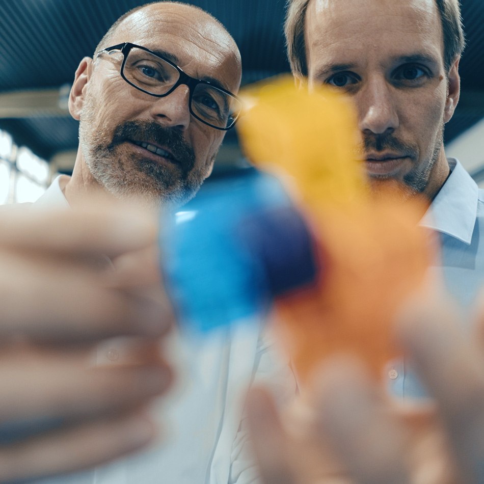 Two engineers are holding two objects together and analysing them. The focus is on the engineers' faces, with the object at the front de-focused.